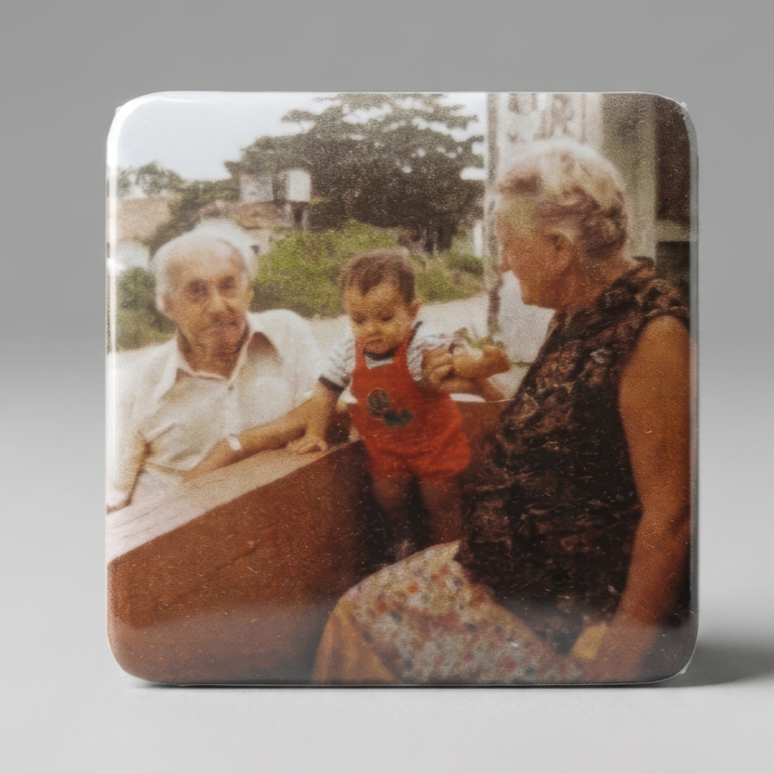 Ímã com foto antiga colorida de bisavós ao lado de bisneto em pé sobre banco de madeira